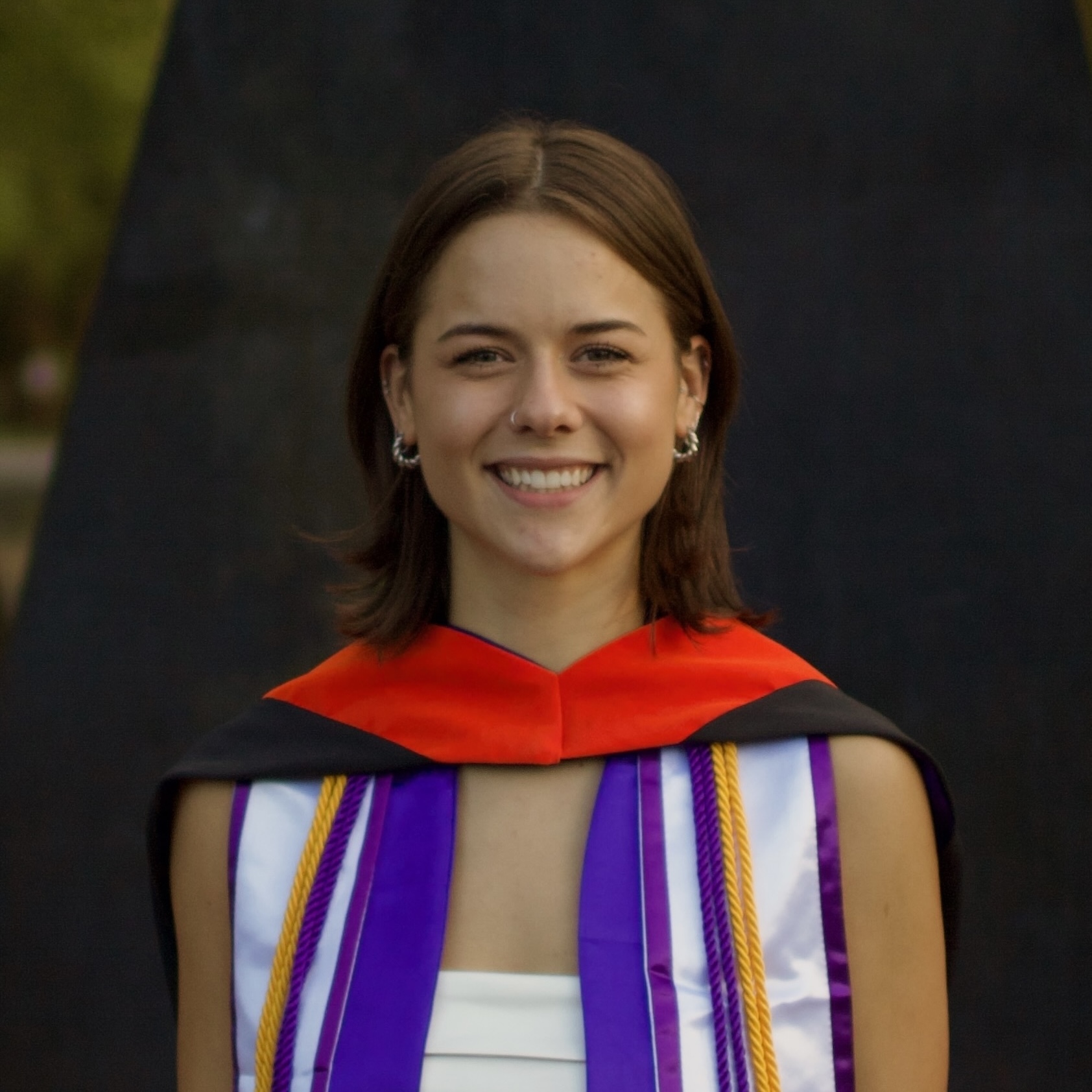 Headshot of a white woman with shoulder-length brown hair, smiling in her graduation regalia and a bright orange master's hood.